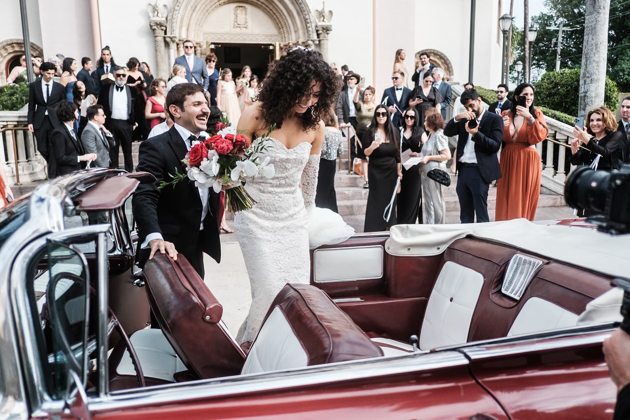 A bride and groom stepping into a red convertible after their church ceremony as guests watch from the steps